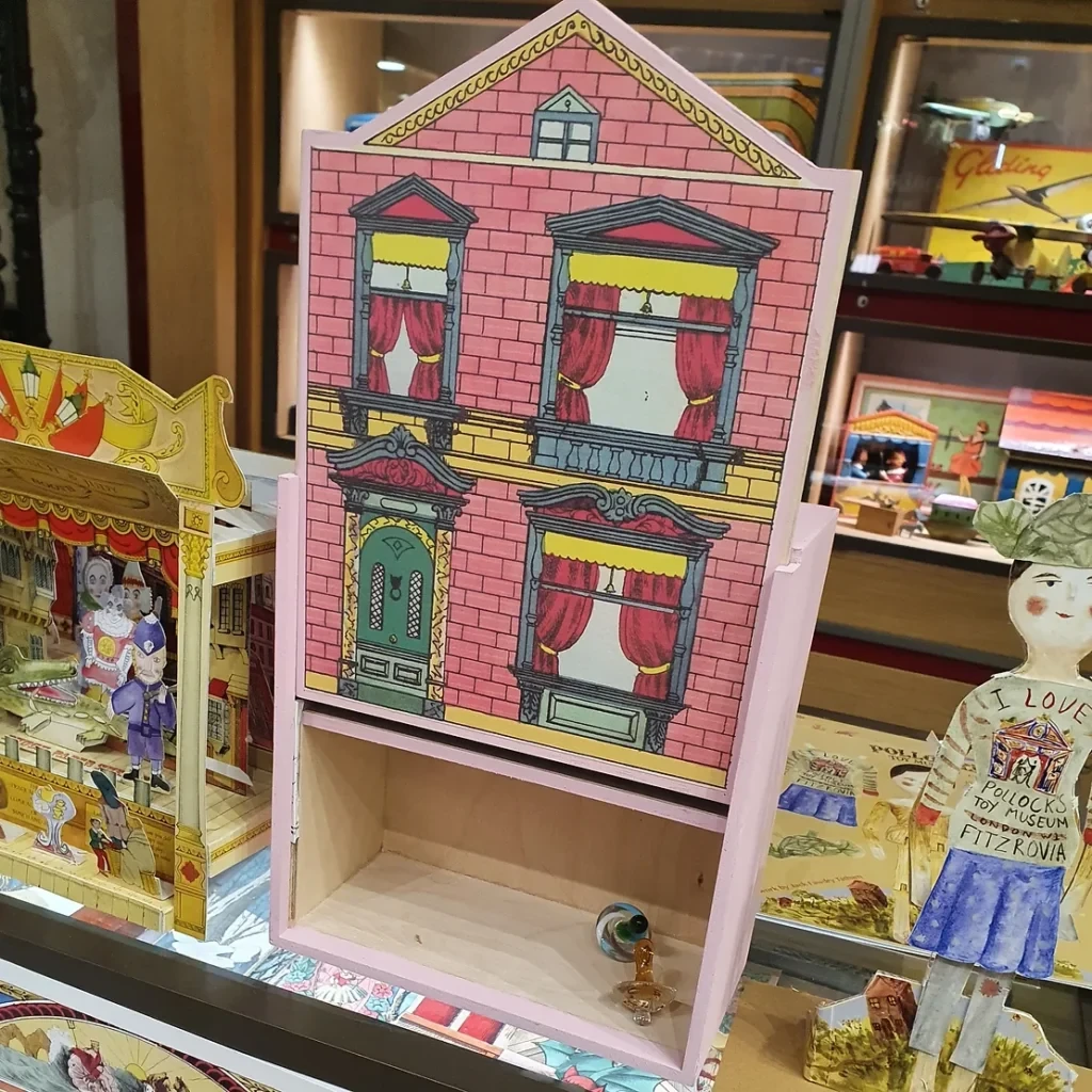 A pink wooden dollhouse with a pretty printed paper facade stands on a shelf next to a paper toy theatre and a paper doll.