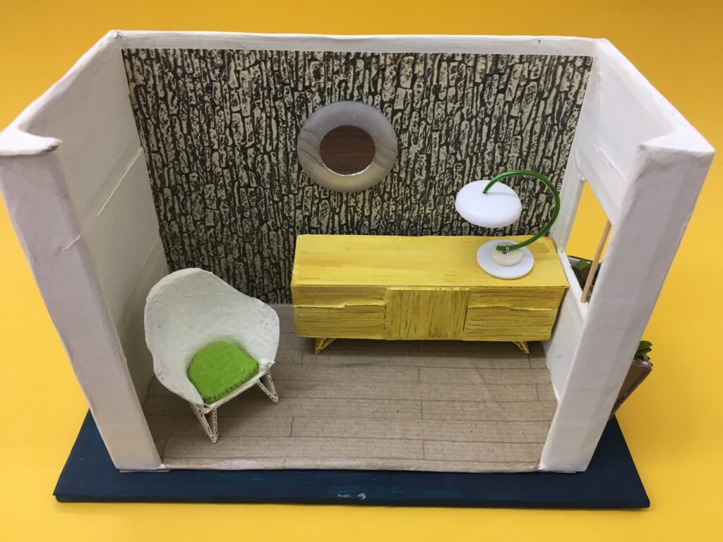 A newly made cardboard miniature room box with white walls, black wood effect wallpaper, a yellow sideboard and white and green bucket chair and lamp on a black base with a mustard background.