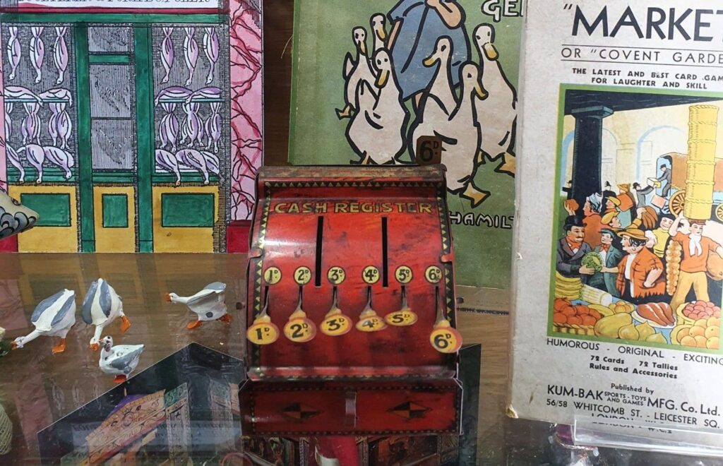 A bright red tin toy cash register sits on a shelf surrounded by children's books and tiny toy geese