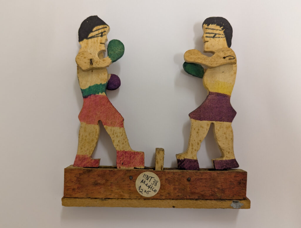 Two brightly painted wooden boxers fighting on a plinth. Their arms are jointed so they can swing a punch.