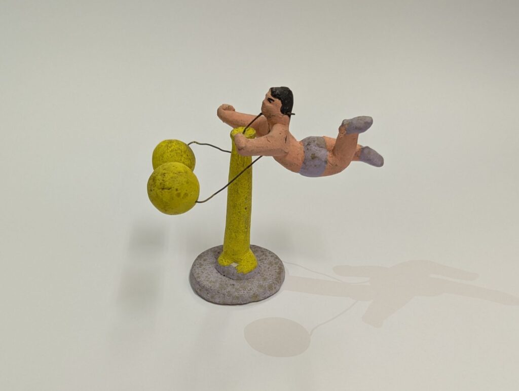 Spinning acrobat balance toy from Mexico, made out of clay. The male acrobat's legs extend out behind him as he spins around a bright yellow pole.