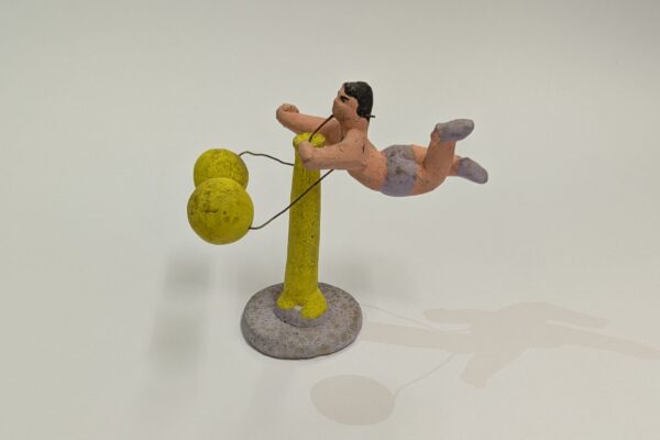 Spinning acrobat balance toy from Mexico, made out of clay. The male acrobat's legs extend out behind him as he spins around a bright yellow pole.