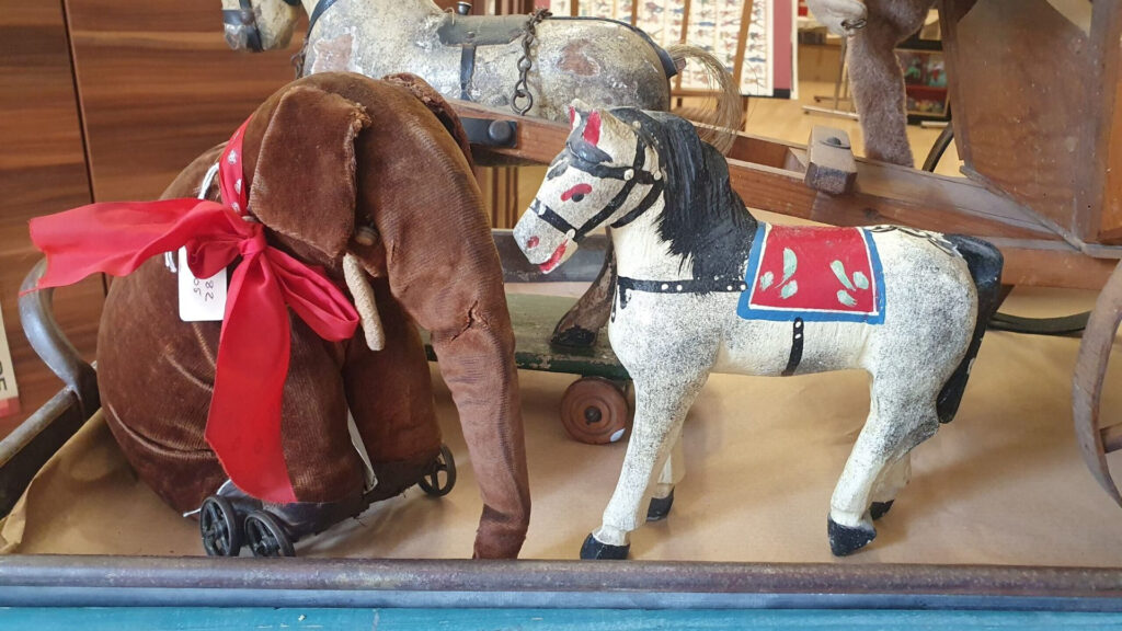 A soft brown velvet elephant on wheels with a big red ribbon and a model horse with painted saddle and reins sit looking at each other. Both are vintage and well loved.