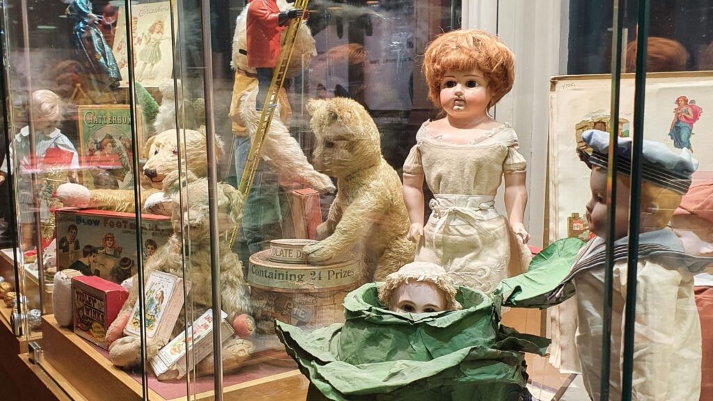 Various antique toys including a doll hiding in a cabbage, teddy bears, a sailor doll, and others, all displayed within a glass cabinet.