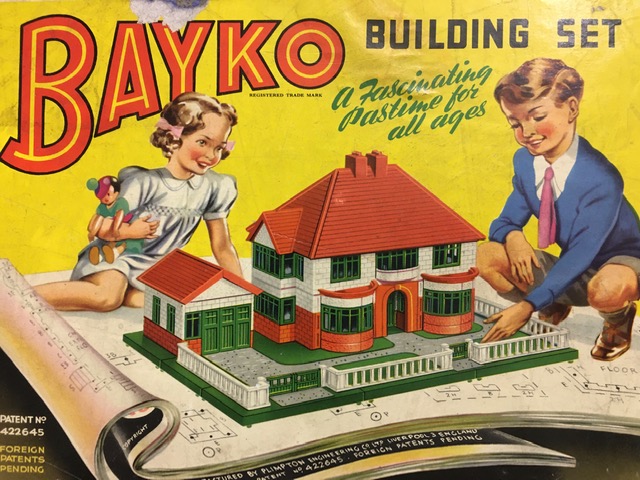Photo of the box art from a vintage Bayko Building Set, "a fascinating pastime for all ages". On the picture a young boy and girl dressed in clothing from the 1950s are admiring a suburban house they have made from the kit.