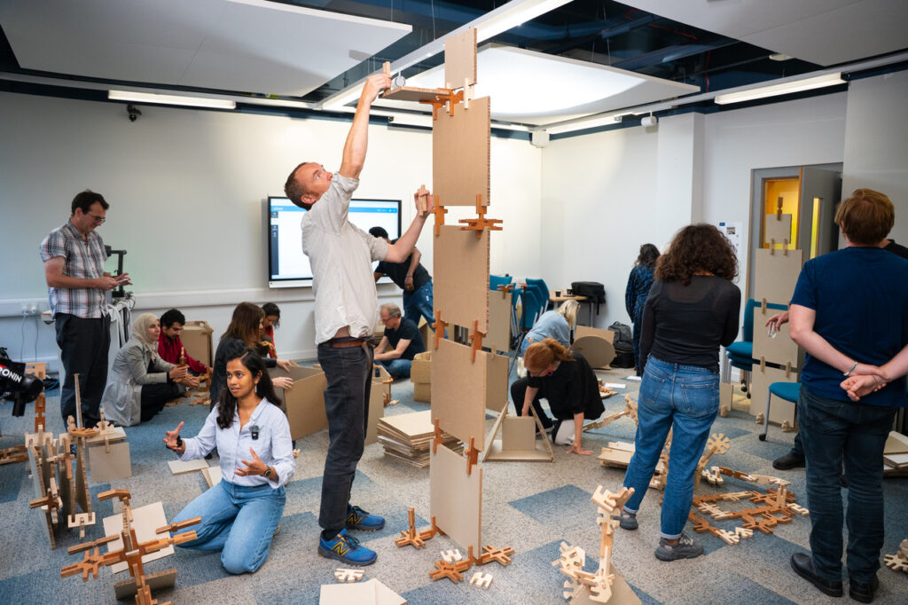 A group of people take part in a workshop in an open-plan room, playing with the giant Mapl construction kit to make abstract structures and models.