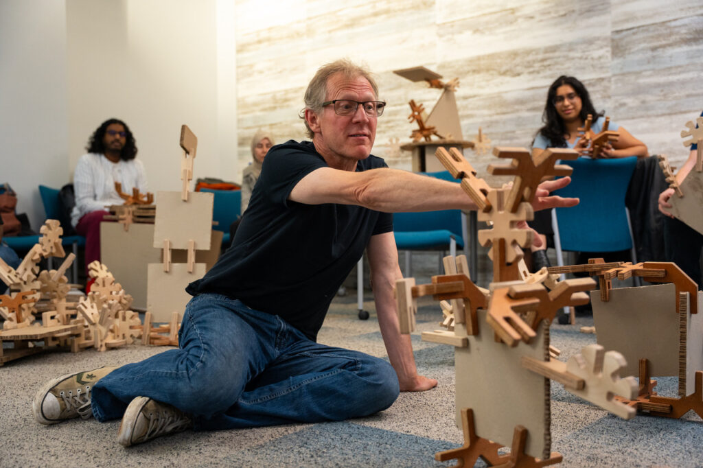 A man sits on the floor reaching out to a large abstract model made up of wooden or cardboard pieces that slot together, from the Mapl construction kit. He is surrounded by other models and workshop participants.
