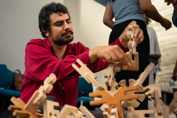 A man works on a Mapl construction kit model with an intense expression of concentration. He is slotting a piece shaped like a circular cog with spokes into another piece that is more angular, to make his creation taller.