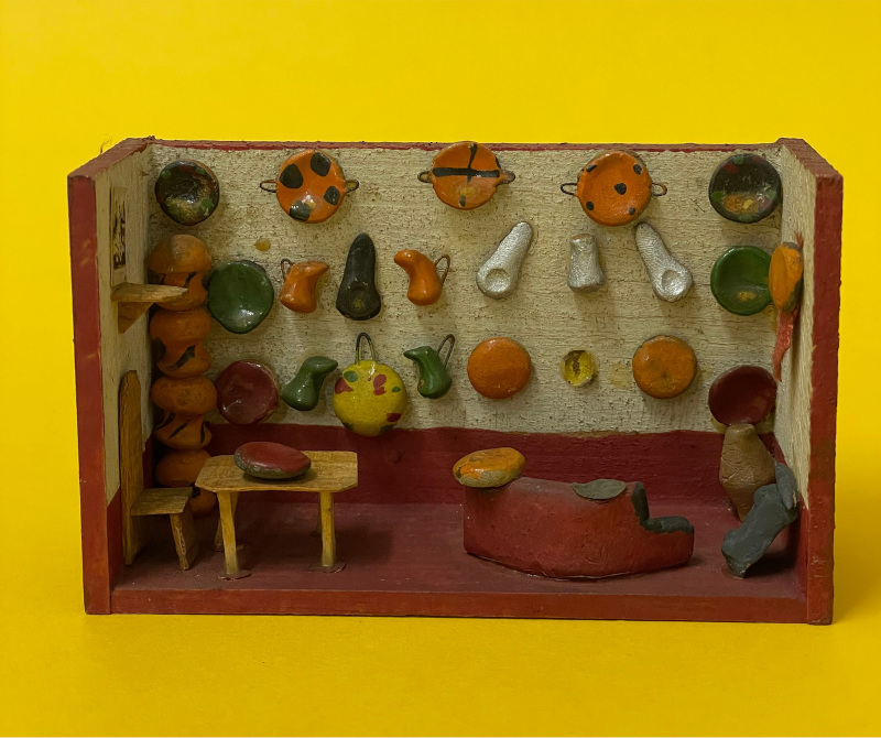 Traditional Mexican miniature diorama from the Pollock's Toy Museum collection, showing a kitchen with table and chair, and a back wall hung with brightly coloured miniature pots, pans and jugs.