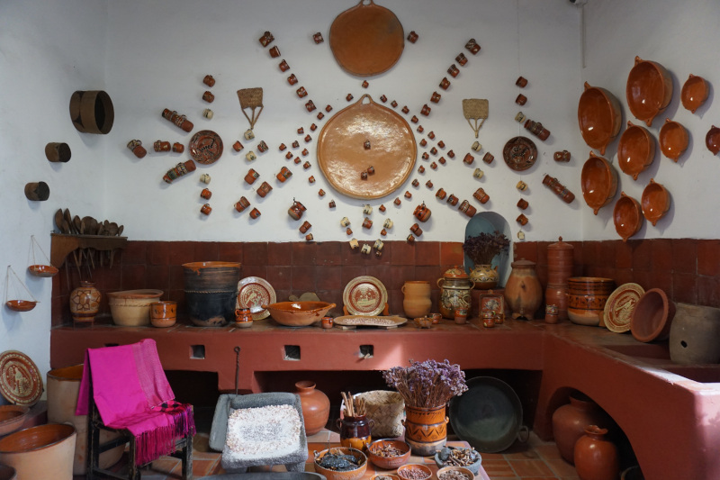 A real-life traditional Mexican kitchen, featuring a large collection of traditional terracotta coloured ceramic mugs, plates and other items, hung in a decorative display on the wall and lined up along the surfaces. Photo credit: Andrearodr, CC BY-SA 4.0, via Wikimedia Commons