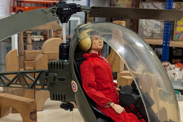 A vintage Action Man figure in a red jumpsuit and white helmet pilots a plastic toy helicopter.