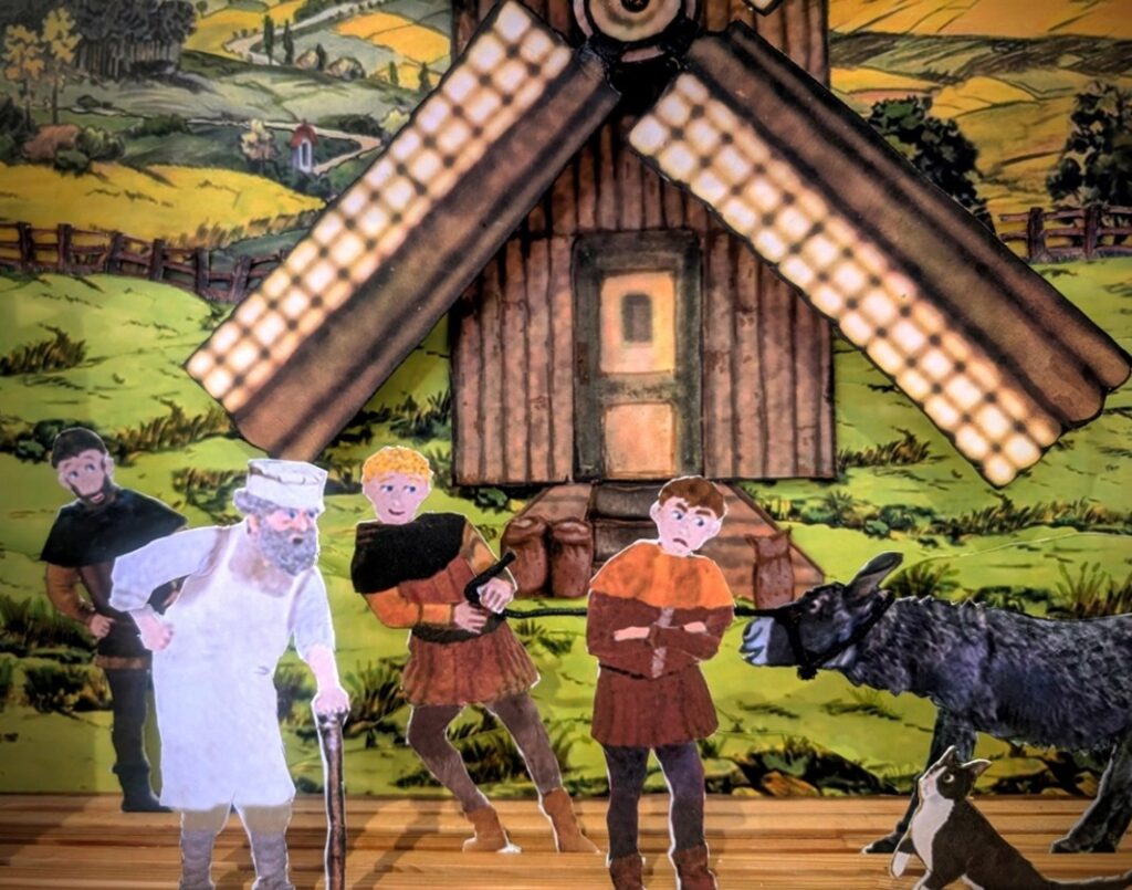 Photograph of a colourful contemporary paper theatre scene, from the story of Puss in Boots, featuring an old man and three younger men, a donkey, and a cat, standing and arguing in front of a windmill.