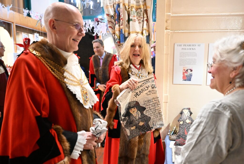 Dame Susan Langley, the Lady Mayor of London, holds up a Pearly Queen doll kit. The Lady Mayor and her entourage are wearing red robes trimmed with fur, and their chains of office. They are talking to Debby Brown, the museum's curator.