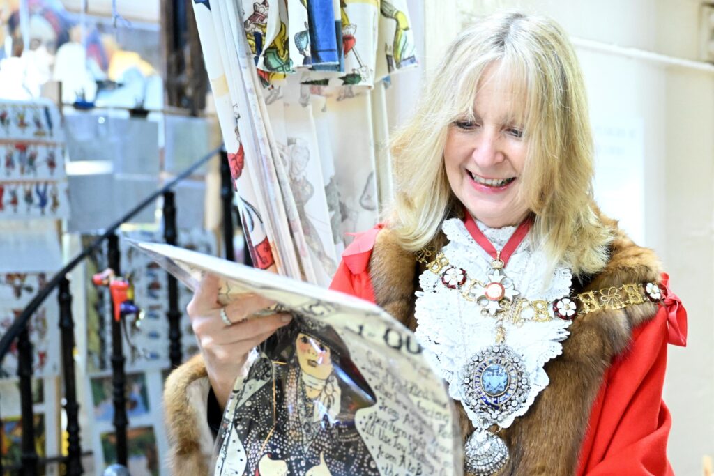 Dame Susan Langley, the Lady Mayor of London, is smiling as she looks at a Pollock's Pearly Queen doll kit, designed by Maria Wood. The Lady Mayor is wearing a red robe trimmed with light brown fur, over which is a white lace collar and an impressive jewelled and enamelled chain of office. The distinctive spiral staircase in the Leadenhall venue can just be seen behind her.