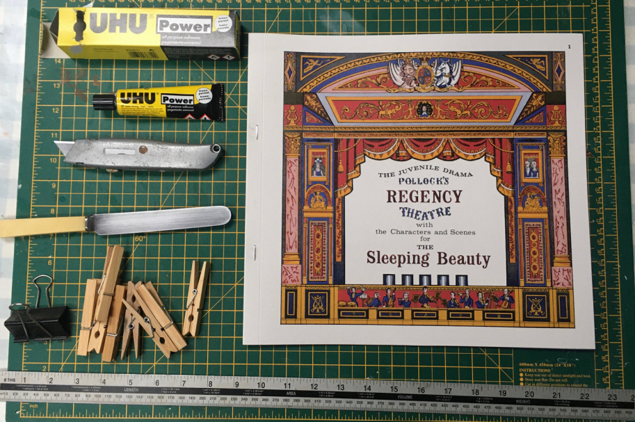 The Pollock's Toy Museum Regency Theatre kit book lies to the right on a green, gridded cutting mat. On the left are various tools set out ready for construction, including glue, a craft knife, a butter knife, and clips. A long metal ruler lies across the bottom of the grid. 