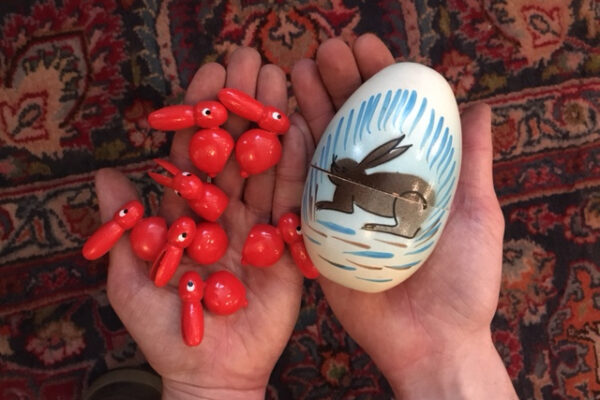 A pair of outstretched hands, palm up, hold out a white toy egg painted with an illustration of a hare, and a number of small red-painted toy hares.