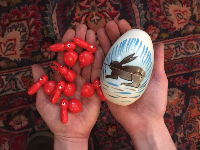 A pair of outstretched hands, palm up, hold out a white toy egg painted with an illustration of a hare, and a number of small red-painted toy hares.