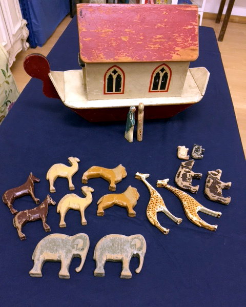 A wooden Noah's ark toy with a red-painted roof and hull stands on a blue tablecloth. Arranged in front of it are carved wooden figures of Noah and his wife, and seven pairs of animals, including elephants, giraffes and lions.  