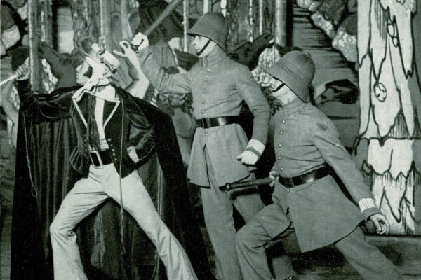 Black and white photograph of three male dancers, two dressed as police officers carrying truncheons, all striking a power pose and looking off to the left, highly alert, on a stage with giant line-drawn toy theatre scenery.