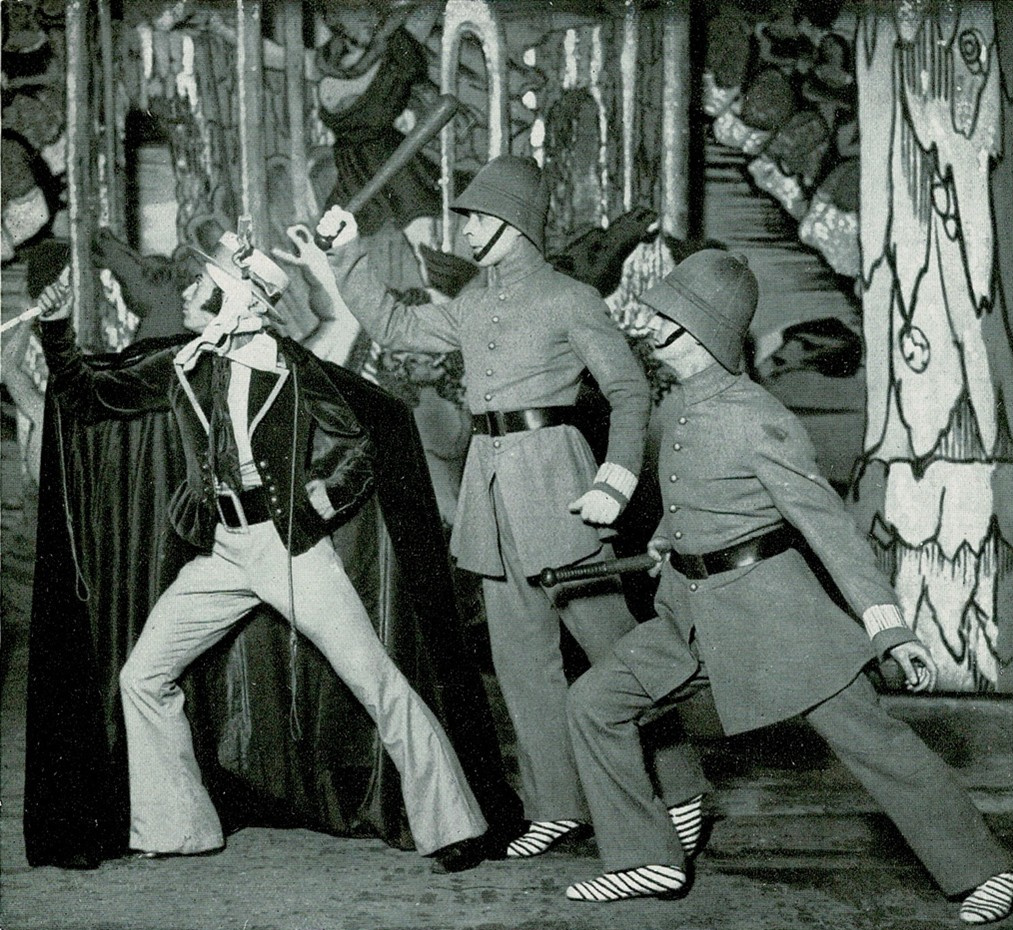 Black and white photograph of three male dancers, two dressed as police officers carrying truncheons, all striking a power pose and looking off to the left, highly alert, on a stage with giant line-drawn toy theatre scenery.