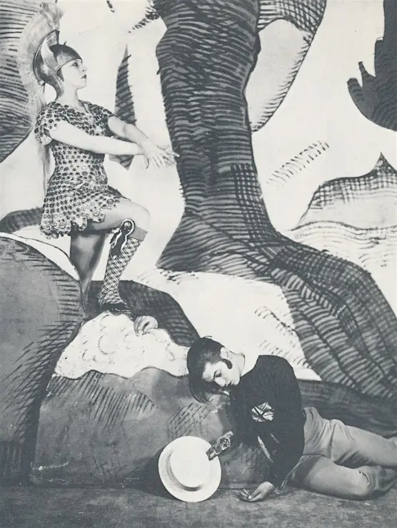 Black and white photograph of two dancers, one woman and one man, strikingly posed in front of scenery enlarged from a toy theatre to giant size. The woman wears a crested helmet and holds her arms in front as if riding a horse, while the man lies on the ground, one arm flung onto the scenic rock, as if exhausted, dead, or in despair.