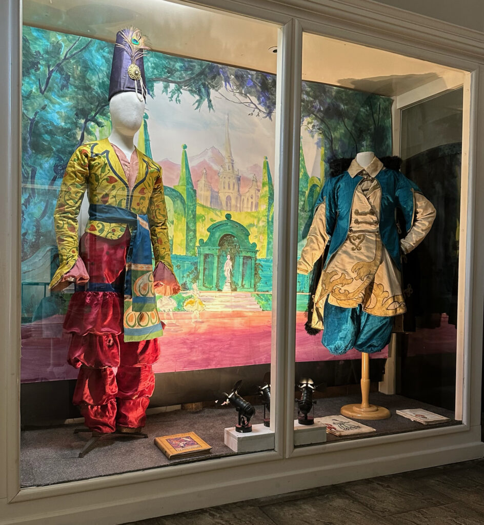 Photograph of two mannequins wearing brightly coloured male dancers' costumes from a ballet, stood in front of a painted scenic backdrop.