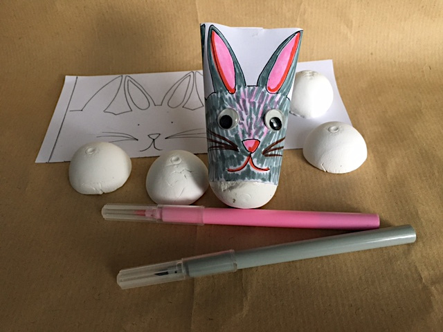 In front of a printed bunny template stands a fully handmade Easter bunny toy, with googly eyes and a wobbly polystyrene-like bottom. Felt tips in pink and grey lie nearby, ready to decorate the next bunny, and there are a number of polystyrene half-spheres ready to wobble more Easter rabbits.